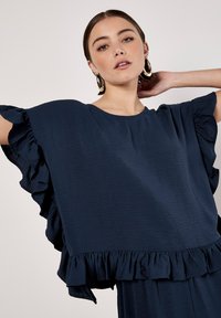 Marineblaues Oberteil aus strukturiertem Stoff, mit Rüschen an den Ärmeln und am Saum, im lockeren Schnitt und mit rundem Ausschnitt.