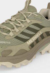 Gros plan d'une chaussure de randonnée Merrell verte avec tissu Gore-Tex, semelle robuste et lacets à motifs sur fond clair.