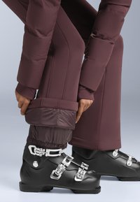 Bordeauxrode skibroek met een gewatteerde textuur, elastische boorden en zwarte skistapschoenen met zilveren gespen en een glanzende afwerking.