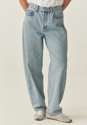 Osoba ubrana w jasnoniebieskie, damskie jeansy z wysokim stanem i luźnym krojem, z rękami w kieszeniach, oraz białe buty sportowe na jednolitym tle.