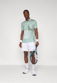 Polo verde menta con motivi geometrici bianchi e verdi, abbinato a pantaloni corti bianchi e sneaker bianchi, mentre tiene una racchetta da tennis.