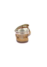 Goldene metallische Sandale mit flacher Sohle und verstellbarem Riemen, mit abgerundetem Zehenbereich und Lederinnensohle, vor einem weißen Hintergrund.