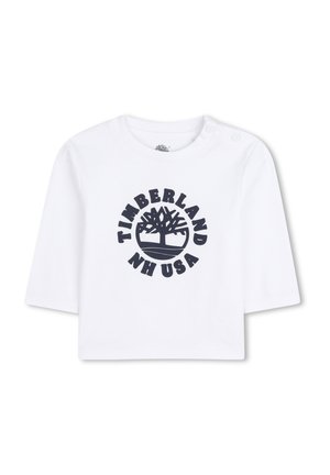 Chemise blanche en coton à manches courtes arborant un logo circulaire Timberland bleu marine avec un graphique d'arbre et le texte "NH USA." Deux boutons-pression aux épaules.