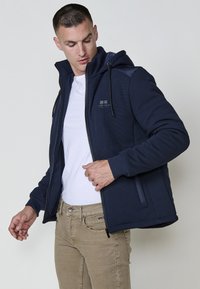 Giacca zip-up blu navy con cappuccio, in tessuto testurizzato, con polsini elastici e pannelli sulle spalle a contrasto. Abbinata con una maglietta bianca e pantaloni beige.