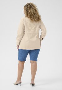 Chaqueta beige de botonadura con bordado de sol en la parte posterior, combinada con pantalones cortos de mezclilla azul a rayas y sandalias de tacón beige.