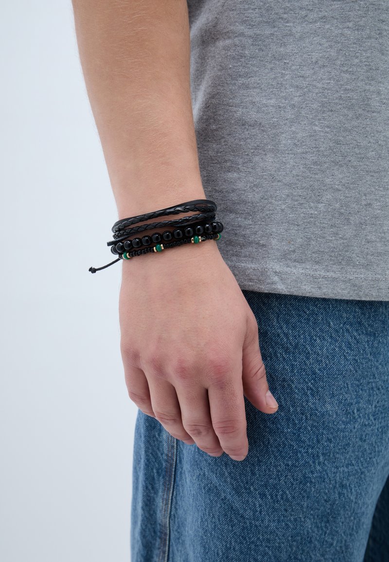 Alkar és kéz három fekete karkötővel, egy fonott bőr és két gyöngyös, kék farmerral és szürke inggel viselve.