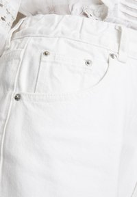Gros plan sur une poche de jean blanc en denim avec des rivets argentés et une ceinture, associé à un haut blanc texturé et en dentelle.