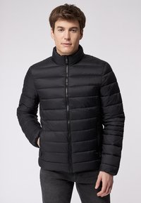 Giacca imbottita nera con colletto alto, chiusura frontale con zip e tasche laterali. Il design presenta sezioni trapuntate orizzontali e una superficie liscia.