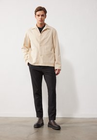 Veste boutonnée beige clair avec un col pointu, deux poches avant, portée sur une chemise noire et associée à un pantalon noir et des boots marron à chevilles.
