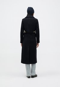 Lang mørkeblå frakke med en struktureret krave, knapper ved taljen og lige snit. Båret over lyse denimjeans og sorte sko.