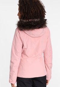 Frau mit lockigem Haar, die eine hellrosa Winterjacke mit einer mit Fell ausgekleideten Kapuze trägt, von hinten vor einem weißen Hintergrund gezeigt.