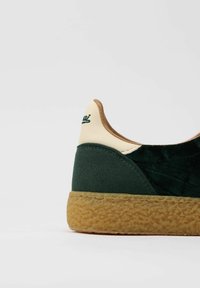 Sneaker verde in velluto con un accento in pelle crema sul tallone, dotata di suola in gomma texturizzata e dettaglio del logo discreto.