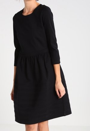 Vrouw draagt een zwarte jurk tot op de knie met lange mouwen, een geplooide taille en getextureerde horizontale strepen vlakbij de zoom.