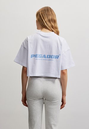 Femme aux longs cheveux portant un T-shirt court blanc avec le texte bleu "PEGADOR" et un pantalon ajusté gris clair, vue de dos.