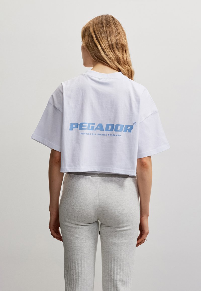 Γυναίκα με μακριά μαλλιά που φοράει άσπρο crop top με μπλε κείμενο "PEGADOR" και ανοιχτό γκρι εφαρμοστά παντελόνια, εμφανιζόμενη από πίσω.