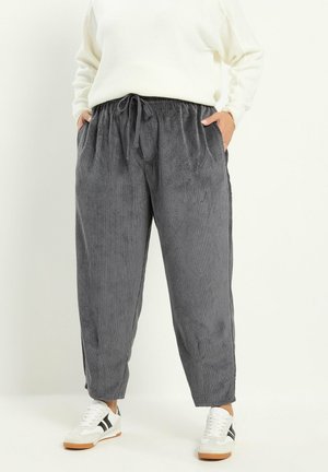 Personne portant un pantalon large en velours côtelé gris avec taille élastique et cordon, un pull blanc et des baskets blanches avec des rayures noires.