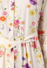 Rochie florală din material ușor, cu un fundal alb, imprimeuri multicolore cu flori, nasturi și detaliu de talie legat.