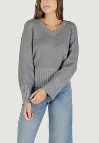 Maglione grigio a V in maglia con texture a coste all'orlo e ai polsini, caratterizzato da maniche ampie. Abbinato a jeans azzurri chiari.