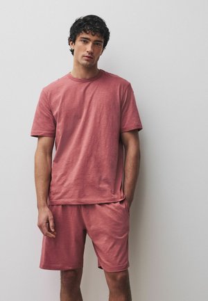 Mann mit dunklen lockigen Haaren, der ein locker sitzendes, staubrosafarbenes T-Shirt mit kurzen Ärmeln und passende Shorts trägt und vor einer schlichten weißen Wand steht.