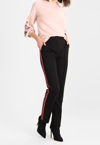 Hellrosa Pullover mit Blumenstickerei an den Ärmeln und schwarze Hose mit roten Seitenstreifen, kombiniert mit schwarzen Stiefeln mit hohem Absatz.