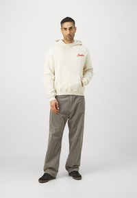 Sudadera crema con bolsillo frontal y logo de texto rojo. Combinada con pantalones de pana grises y zapatillas negras. Diseño simple y corte relajado.