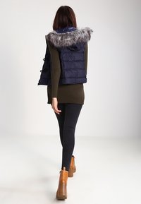 Marinblå pufferväst med en fuskpälsluva, kviltat mönster, bärs över en olivgrön tröja, matchat med svarta leggings och sandfärgade ankelstövlar.