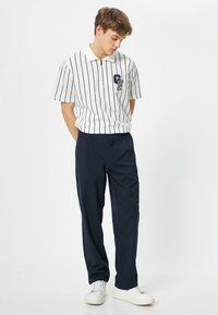 Valkoinen raidallinen polo-paita, jossa on kaulus ja rintalogot, yhdistettynä tummansinisiin housuihin, joissa on rento leikkaus ja sivutaskut. Valkoiset lenkkarit.