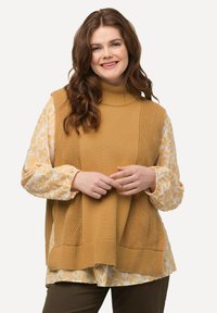 Sweter z golfem w kolorze musztardowym, zestawiony z jasną bluzką w kwiaty z bufkami. Modelka stoi z dłońmi złożonymi.