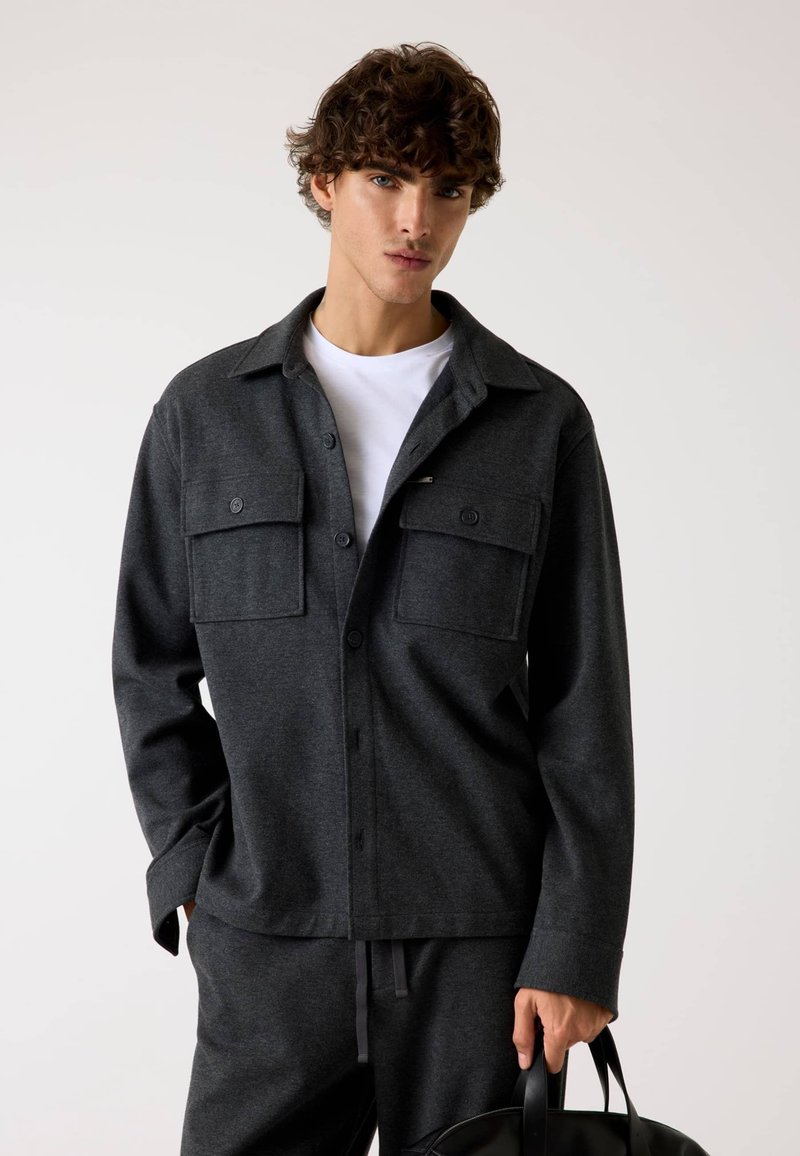 Camicetta button-up grigio scuro realizzata in un tessuto morbido, dotata di due tasche sul petto, colletto e vestibilità comoda. Il modello tiene una borsa nera.