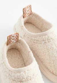 Luddiga beige slip-on skor med en mjuk, texturerad yta. Har vävda rosa och vita detaljer vid hälen och en gummisul.