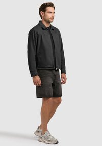 Hombre con chaqueta negra de cremallera, pantalones cortos de mezclilla oscura y zapatillas grises New Balance, de pie contra un fondo gris claro.