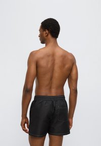 Μαύρο μαγιό σόρτς με ελαστική μέση και πίσω τσέπη, με λευκή ραφή. Το μοντέλο φαίνεται από πίσω.