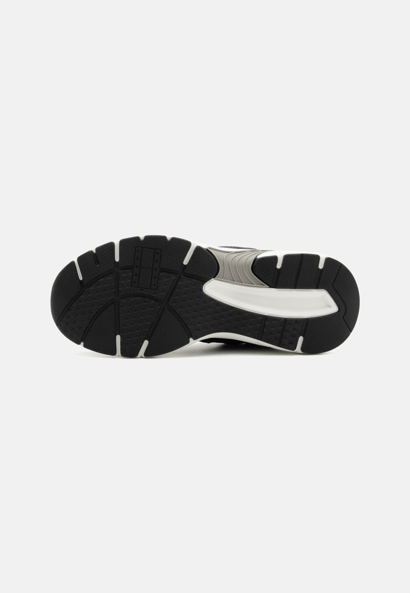 Semelle de chaussure de sport noire, avec du caoutchouc texturé, des rainures motifées et des décorations blanches sur les côtés, offrant adhérence et stabilité.