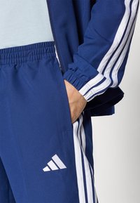 Μπλε αθλητική φόρμα με λευκές πλευρικές ρίγες και με κεντημένο λογότυπο. Απαλή ύφανση με ελαστική μέση και μανσέτες.