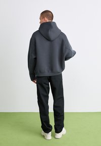 Donkergrijze hoodie met een ruime fit en een grote capuchon, gecombineerd met zwarte broek en witte sneakers, op een groen tapijt.