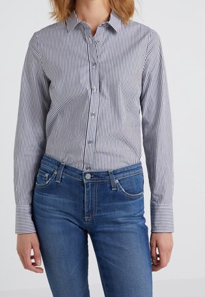 Frau trägt ein blau-weiß gestreiftes, langärmliges Hemd mit Knöpfen, das in eine blaue Jeans gesteckt ist, und steht vor einem schlichten Hintergrund.