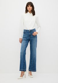 Szerokie niebieskie dżinsy z perłowymi zdobieniami, wysoki stan, postrzępione nogawki, w zestawieniu z dopasowanym białym topem z długim rękawem i beżowymi szpilkami.