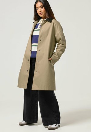 Kobieta w beżowym płaszczu trenczowym na wierzch paskowanego swetra, czarne szerokie spodnie i trampki, pozuje z jedną ręką w kieszeni płaszcza.
