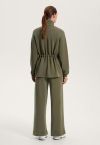 Olivno zelena pulover z visokim ovratnikom in vezico v pasu, v kombinaciji z širokimi hlačami. Gladka tekstura tkanine, sproščen silhuet.