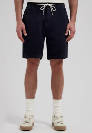 Man die zwarte corduroy short met wit trekkoord, witte sokken en witte sneakers draagt, staande tegen een grijze achtergrond.