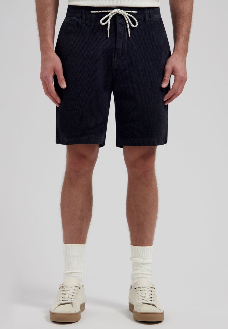 Homme portant un short en velours côtelé noir avec un cordon blanc, des chaussettes blanches et des baskets blanches, debout devant un fond gris.