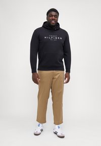 Sudadera negra con capucha y bolsillo frontal, con un logo en texto blanco, combinada con chinos beige y zapatillas blancas con acentos oscuros.