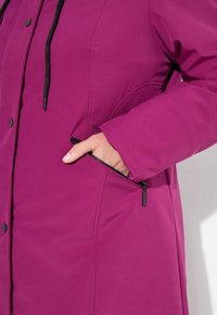Hand in pocket of zipped magenta winter coat with black drawstring and snap buttons, showing coat's fabric texture and stitching details.