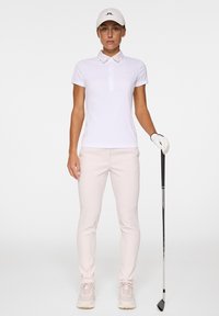 Biała koszulka golfowa z kołnierzykiem, połączona z jasnoróżowymi spodniami. Strój uzupełnia biała czapka i kij golfowy.