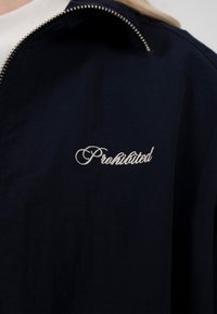Veste marine avec fermeture éclair argentée partiellement ouverte, mot brodé "Prohibited" en cursive blanche à l'avant.