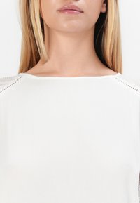 Femme avec de longs cheveux blonds portant un haut texturé blanc avec un col rond, vue des épaules vers le haut contre un fond uni.