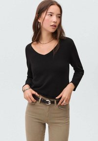 Črna dolgorasna V-izrez pulover iz mehkega materiala, oblečen z oprijetimi bež kavbojkami in črnim pasom, dopolnjen z srebrnimi nakitnimi akcenti.