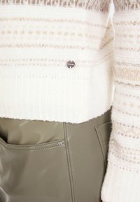 Leicht strukturiertes weißes Sweater mit beigen Streifen und Pailletten, kombiniert mit olivgrünen, kunstledernen Hosen mit sichtbarem Logo.