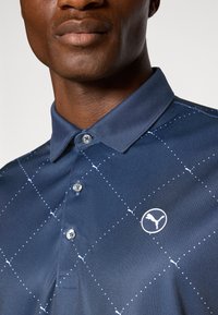 Polo bleu marine avec un motif en losange et des accents blancs. Il dispose d'un col, d'une fermeture à boutons et d'un logo sur la poitrine gauche.