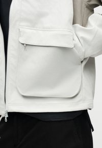 Κοντινό πλάνο ατόμου που φοράει ανοιχτό μπεζ μπουφάν με μεγάλο μπροστινό τσέπη και φερμουάρ, σε συνδυασμό με μαύρο παντελόνι.